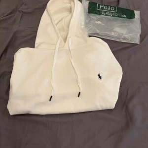 Vit hoodie från Polo Ralph Lauren - Vit hoodie från Polo Ralph Lauren med klassisk svart logga broderad på bröstet. Tröjan har huva med snörning och en stor magficka. Perfekt för dig som gillar stilren och enkel design. Det står att storleken är L men den passar mig och jag har S så köp om du har S så kommer den sitta perfekt.