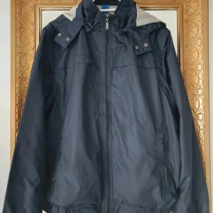 Mörkblå vindjacka med huva - Snygg mörkblå vindjacka med huva och grått innerfoder. Jackan har dragkedja framtill, praktiska fickor med dragkedja och elastisk nederkant. Perfekt för blåsiga dagar och enkel att matcha med olika outfits.