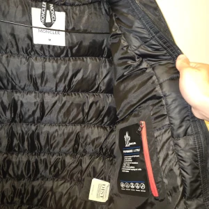 Svart Moncler pufferjacka med huva - Svart pufferjacka från Moncler med huva och dragkedja framtill. Jackan har breda, vadderade kanaler och stora Moncler-loggor på insidan av huvan och längs insidan. Två fickor med dragkedja och klassisk Moncler-patch på ärmen. Perfekt för kalla dagar.