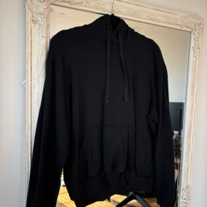 Svart hoodie med huva och ficka - En klassisk svart hoodie med huva och snörning samt en stor magficka. Tröjan har långa ärmar och ribbade muddar vid ärmslut och nederkant. Perfekt för en chill och avslappnad stil.