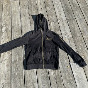 Svart hoodie från BOSS med guld-detaljer - Svart hoodie från BOSS med guldiga dragkedjor och logga på bröstet. Tröjan har huva med BOSS-tryck, långa ärmar och ribbade muddar. Perfekt för dig som gillar stilrena och exklusiva streetwear-plagg.