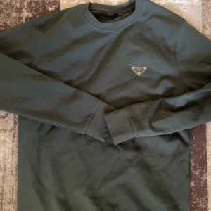Mörkgrön sweatshirt från Prada - Snygg mörkgrön sweatshirt från Prada med klassisk rund hals och diskret logga på bröstet. Tröjan har långa ärmar och är tillverkad i mjuk bomull som känns skön mot huden. Perfekt för dig som gillar stilrena designer och exklusiva märken.