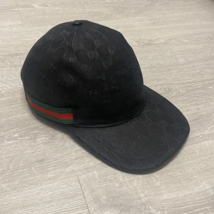 Gucci keps  - (Ny färgad) Snygg Gucci keps med diskret GG-monogram över hela ytan. Klassisk böjd skärm och justerbar rem baktill. Sidan pryds av Guccis ikoniska gröna och röda rand. Perfekt accessoar för en trendig streetstyle-look.