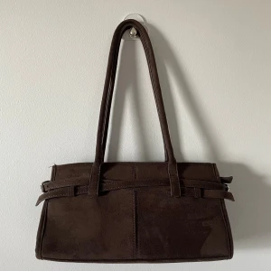 Brun mocka axelväska med bältdetalj - Stilren brun axelväska i mocka med långa handtag och snygga bältdetaljer på sidorna. Använd 2 gånger. Prisen kan diskuteras 