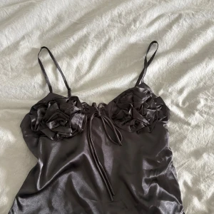 Svart satin linnetopp med rosetter - Säljer en svart satin linnetopp med smala axelband och dekorativa rosetter framtill. Toppen har en glansig finish och vackra volangdetaljer över bysten som ger en romantisk vibe. Perfekt för dig som gillar unika detaljer och en feminin look. Storlek M