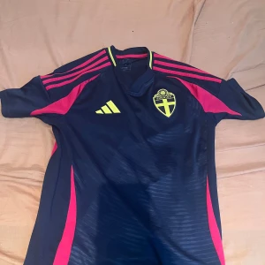 Sverige fotbollströja Adidas marinblå S - Svensk landslagströja från Adidas i marinblått med neon-gula detaljer och rosa ränder på axlarna. Broderad Sverige-logga på bröstet och tryckt text i nacken. Tillverkad i lätt och ventilerande funktionsmaterial, perfekt för match eller träning.