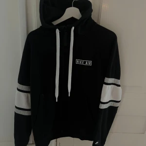 Svart Nike Air hoodie med vita detaljer - Nike Air hoodie i svart med vita snören och vita ränder på ärmarna. Dragkedja framtill och huva för extra streetstyle. Snyggt Nike Air-tryck på bröstet och fickor på sidorna. Perfekt för chill eller häng med kompisar. (Oversized) i passformen.