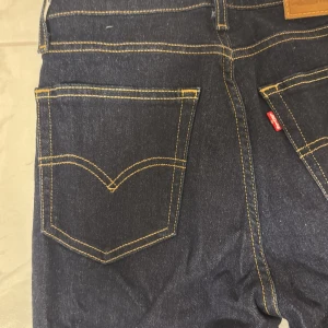 Levi's 501 mörkblå jeans straight fit - Klassiska Levi's 501 jeans i mörkblå denim med raka ben och kontrastsömmar. Byxorna har fem fickor, läderpatch bak och den ikoniska röda Levi's-taggen. Perfekta för dig som gillar tidlös stil och vill ha ett par jeans som funkar till allt.