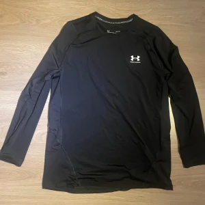 Svart långärmad träningströja UA - Svart långärmad träningströja från Under Armour med vitt logotryck på bröstet. Tillverkad i ett lätt och stretchigt syntetmaterial, perfekt för intensiva träningspass. Snygg och sportig passform som sitter tajt mot kroppen.