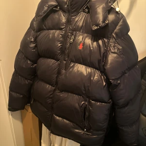 Svart pufferjacka Polo Ralph Lauren XL - Säljer en svart pufferjacka från Polo Ralph Lauren i storlek XL men passar även M. Jackan har glansig finish, klassisk röd Polo-logga på bröstet, flera dragkedjefickor och hög krage. Perfekt för kalla dagar med sin tjocka vaddering och sportiga look.