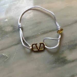 Valentino armband med vit snodd - Snyggt armband från Valentino med justerbar vit snodd och guldfärgat V-logo i metall. Enkelt och stilrent smycke som ger en lyxig touch till din outfit. Perfekt för dig som gillar designeraccessoarer och vill ha något diskret men ändå exklusivt.