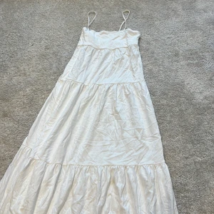 Vit långklänning från H&M - Superfin vit långklänning från H&M med tunna axelband och volangiga lager. Klänningen har en luftig och flowy passform, perfekt för varma dagar. Tillverkad i mjuk bomull som känns skön mot huden. Enkel och stilren design som är lätt att matcha.