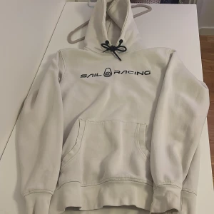 Vit hoodie från Sail Racing med tryck - Vit hoodie från Sail Racing med svart logotyptryck på bröstet  Tröjan har huva med svarta snören och en stor magficka. Mjuk insida och klassisk passform, perfekt för chill dagar.