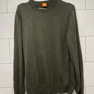 Olivgrön stickad tröja från Hugo Boss - Snygg olivgrön stickad tröja från Hugo Boss med rund halsringning och diskret logga vid nederkanten. Tröjan har lång ärm och klassisk passform, perfekt för lager-på-lager. Materialet känns mjukt och ger en clean look. Hör av dig vid minsta fråga. Mvh