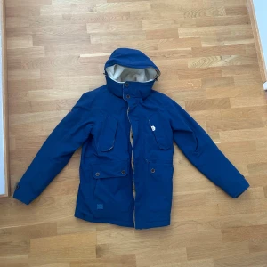 Blå jacka från Didriksons - Snygg blå parkasjacka från Didriksons med mysigt vitt teddyfoder och röda detaljer. Jackan har flera stora fickor med knappar, huva och dragkedja framtill. Perfekt för kalla dagar med sin varma insida och praktiska design.