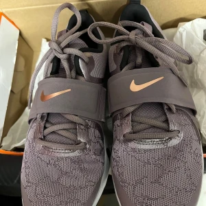 Nike React sneakers i lila mesh - Nike React sneakers i en snygg lila nyans med mesh-ovandel och breda elastiska band över foten. Skorna har kopparfärgade swoosh-loggor, vit och svart sula med React-dämpning och coola detaljer. Perfekta för dig som vill ha både stil och komfort.