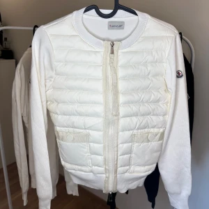 Moncler Cardigan - Snygg vit cardigan jacka från Moncler med quiltad framsida och stickade ärmar. Jackan har dragkedja framtill, rund hals och två fickor med knappstängning. Moncler-logga på ärmen ger extra stilpoäng. Perfekt för dig som gillar clean och exklusiv look.