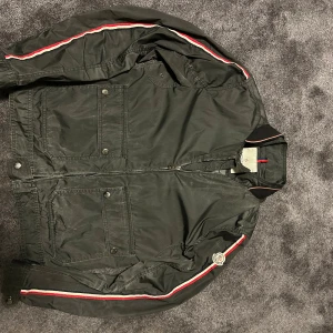 Grå/olivgrön vindjacka från Moncler - Grå/olivgrön vindjacka från Moncler med klassisk passform. Jackan har dragkedja framtill, två stora fickor med knappar och en röd-vit-blå rand längs ärmarna. Materialet är lätt och syntetiskt, perfekt för blåsiga dagar. Moncler-logga på ena ärmen ger en exklusiv touch. skick är 4/5 så de är ganska bra skick, äktehetscertifikat följer med. 