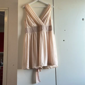 Champagnefärgad kort klänning Xoul Edition - Superfin kort klänning från Xoul Edition i champagnefärgad satinliknande polyester. Djup v-ringning både fram och bak, bred midja med knytband och lätt utställd kjol. Ärmlös och perfekt för sommarens tillfällen. Snyggt fall och elegant look.