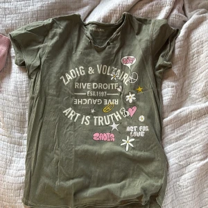 Grön Zadig & Voltaire t-shirt med tryck - Cool grön t-shirt från Zadig & Voltaire med tryck och patchar framtill, bland annat blommor, stjärnor och text. Rund halsringning med två knappar och råa kanter. Perfekt för en avslappnad och trendig look. Köp direkt för 200