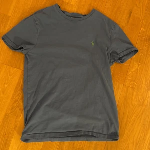Blå t-shirt från Polo Ralph Lauren S - Blå t-shirt från Polo Ralph Lauren i bomull med klassisk rund hals och grön broderad logga på bröstet. Modellen är custom slim fit och har korta ärmar. Perfekt för en clean och stilren look. Säljer den här Polo tröjan då den inte passar mig längre.