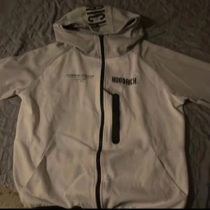 Vit hoodie från Hoodrich med tryck - Vit hoodie från Hoodrich med svart tryck på bröstet, ryggen och huvan. Tröjan har hel dragkedja, huva och en svart detaljficka på bröstet. Tillverkad i mjuk bomullsmix, perfekt för en streetwear-look.