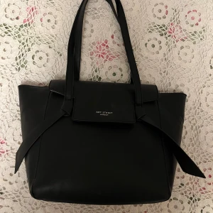 Svart axelväska från Art Street London - Stilren svart axelväska i skinnimitation från Art Street London. Väskan har långa handtag, dekorativa knytband på sidorna och ett innerfoder med prickigt mönster. Praktisk dragkedja baktill och flera fack inuti för smart förvaring.