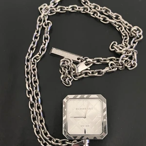 Burberry silverfärgad fickur - Säljer ett stilrent fickur från Burberry i silverfärgat metall med kedja. Klockan har fyrkantig urtavla med graverat mönster och diskreta index. Klassisk Burberry-logga och Swiss Made på urtavlan. Perfekt accessoar för dig som gillar lyxiga detaljer.