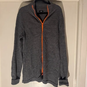 Grå kofta från Mads Nørgaard - Grå stickad kofta från Mads Nørgaard med orange dragkedja framtill. Koftan har hög krage och långärmad design, perfekt för lager-på-lager. Tillverkad i Danmark och har en snygg, diskret struktur i materialet.