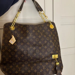  axelväska - Snygg axelväska i klassiskt monogrammönster. Väskan är brun med guldiga metalldetaljer och kedjor, tillverkad i skinn. Rymligt fack med dragkedja och innerfickor, perfekt för dig som vill ha både stil och funktion.