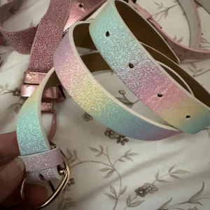 Glittrigt pastellfärgat bälte - Säljer ett bälte i regnbågsfärgade pasteller med glittrig yta och guldfärgat spänne. Bältet har en mjuk övergång mellan ljusblått, rosa, gult och grönt och ger en riktigt drömmig vibe. Perfekt accessoar för att lyfta vilken outfit som helst.