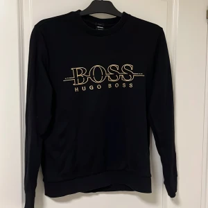 Svart slim fit tröja från Hugo Boss - Svart långärmad tröja från Hugo Boss med stilren BOSS-logga i guld på bröstet. Slim fit-modell med rund halsringning och mjukt material i bomull och polyester. Perfekt för dig som gillar en clean och snygg look.