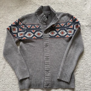 Cardigan - Säljer en grå stickad cardigan med snyggt geometriskt mönster i blått, vitt och orange över axlar och bröst. Koftan har ribbad krage, knäppning framtill och långa ärmar. Perfekt för kyliga dagar!