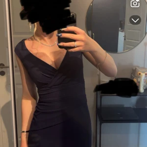 Marinblå långklänning med omlott - Säljer en elegant marinblå långklänning med omlott-design och djup v-ringning. Klänningen är figurnära med breda axelband och har en snygg drapering över midjan. Perfekt för bal eller student. Materialet känns mjukt och följsamt mot kroppen. Är använd bara en gång 