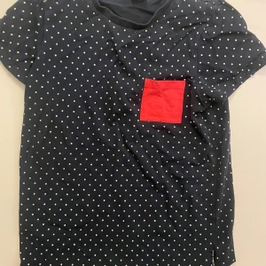 Mörkblå prickig t-shirt Polarn O. Pyret - Snygg mörkblå t-shirt från Polarn O. Pyret med vita prickar över hela och en röd fyrkantig bröstficka som sticker ut. Klassisk rund hals och korta ärmar. Tillverkad i mjuk bomull som är skön mot huden. Perfekt för dig som gillar färg och mönster.