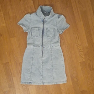 Ljus jeansklänning med dragkedja - Lindex barn - Ljusblå jeansklänning från Lindex med korta ärmar, hög krage och två bröstfickor. Dragkedja framtill ger en cool touch. Perfekt om du vill ge ditt barn en enkel och gullig look.