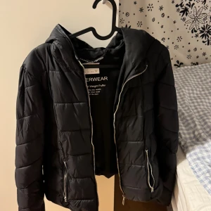 Svart pufferjacka från Bershka - Svart pufferjacka från Bershka med huva och silvriga dragkedjor. Jackan är quiltad och har två fickor med dragkedja framtill. Perfekt för kyliga dagar och har en modern, boxig passform.