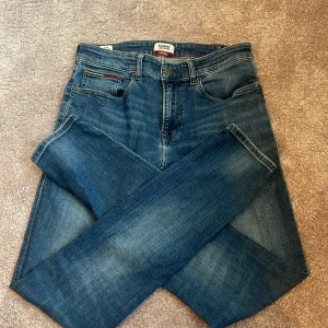 Tommy Jeans Steve Slim Tapered 30/32 - Snygga blå jeans från Tommy Jeans, modell Steve Slim Tapered. Jeansen har klassisk femficksdesign, röda sömdetaljer och en mörk tvätt med lätt slitning. Passformen är slim med avsmalnande ben och materialet är mjukt denim. Perfekt för dig som gillar stilrena jeans med modern siluett.