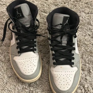 Nike Air Jordan 1 High grå/vit - Säljer ett par Nike Air Jordan 1 High sneakers i grått och vitt med svarta detaljer och snörning. Skorna har en klassisk rund tå, platt sula och är tillverkade i skinn. Swoosh-loggan syns tydligt på sidan och Jordan Wings-märket på ankeln. Perfekt för dig som gillar streetwear. De är äkta 