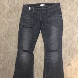 Gråa trendiga bootcut jeans med slitningar - Snygga svarta jeans med bootcut passform och slitna detaljer vid knäna. Jeansen har två framfickor med markerade sömmar och två bakfickor. Dubbel knappstängning i midjan och cool tvättad look. Perfekta för dig som gillar en edgy stil.