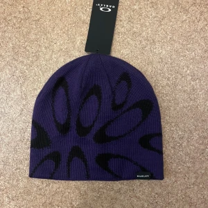 Lila Oakley mössa ellipse graphic - Snygg lila mössa från Oakley med svart grafiskt ellipsmönster. Modellen heter Ellipse Graphic Beanie i färgen Deep Amethyst. Tillverkad i mjukt stickat material och har en liten Oakley-logga framtill. Perfekt för att ge din look en sportig vibe.