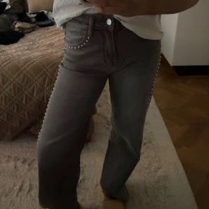Grå wide jeans med pärldetaljer - Snygga grå jeans med breda ben och pärldetaljer längs fickorna och sidorna. Modellen har en klassisk femficksdesign och normal midja. Perfekta för dig som vill sticka ut med coola detaljer och samtidigt ha en bekväm passform.