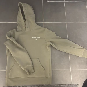 Olivgrön hoodie från Björn Borg - Snygg olivgrön hoodie från Björn Borg med vit logotyp på bröstet. Tröjan har huva med metallöljetter, klassisk känguruficka och långa ärmar med ribbade muddar. Mjuk och skön polyesterblandning som passar perfekt till chill eller träning.