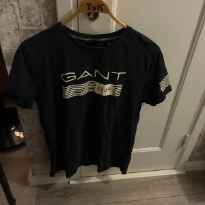 Svart GANT t-shirt med tryck - Snygg svart t-shirt från GANT med vit logga och vågigt tryck framtill samt årtalet 1949. Klassisk rund hals och korta ärmar. Perfekt för en clean och stilren look.