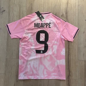Real Madrid Mbappé rosa matchtröja S - Rosa Real Madrid fotbollströja från Adidas med Mbappé och nummer 9 på ryggen. Tröjan har svarta detaljer, klubbmärke på bröstet och 'Emirates Fly Better' tryckt framtill. Tillverkad i lätt och ventilerande Aeroready-material. Slim fit och kortärmad.