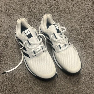 Vita Adidas inomhus skor - Snygga vita Adidas sneakers med klassiska svarta ränder på sidorna. Skorna har snörning, platt sula och är tillverkade i syntetmaterial med textilinslag. Perfekta för dig som gillar sportig och clean stil. Sulan har ett coolt mönster och ger bra grepp. Helt oanvänd pågrund fel storlek