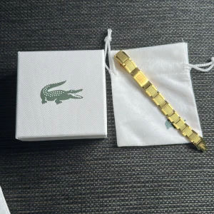 Guldigt armband från Lacoste - Snyggt guldigt armband från Lacoste med länkdesign och detaljerad struktur på varje länk. Armbandet har den klassiska krokodil-loggan ingraverad på spännet och kommer i original ask och tygpåse. Perfekt accessoar för dig som gillar stilrena och exklusiva smycken.