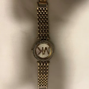 Guldig Michael Kors klocka med stenar - Säljer en snygg guldig klocka från Michael Kors med MK-logga på urtavlan. Klockan har ett armband i metall med flera länkar och glittrande stenar runt boetten. Urtavlan är rund och har en stilren design som sticker ut. Är använd så några stenar fattas därmed pris. Finns ingen låda då det slängdes i flyt