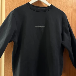 Svart sweatshirt från Calvin Klein Jeans - Svart sweatshirt från Calvin Klein Jeans med diskret logga broderad på bröstet. Tröjan har rund hals, ribbade muddar och lång ärm. Perfekt för dig som gillar stilrena och klassiska plagg med en modern touch. 