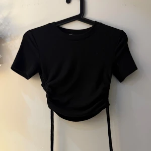 Svart ribbad croppad topp från Zara - Svart kortärmad topp från Zara i ribbad bomull med snörning på sidorna för en snygg croppad look. Rund halsringning och tight passform som ger en cool vibe. Perfekt att styla med jeans eller kjol för en trendig outfit.